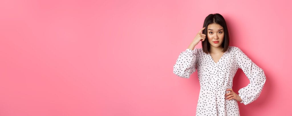 Image of attractive asian lady scolding person, pointing at head and staring at camera disappointed, are you stupid gesture, standing over pink background