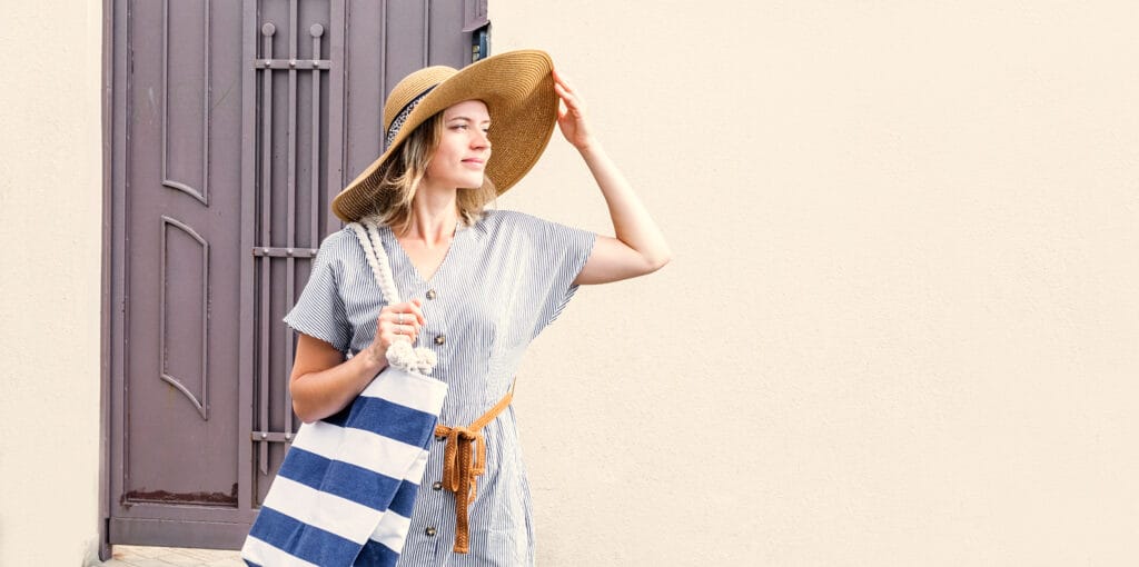 Young blond caucasian woman in summer clothes and hat standing outdoors in the street looking to the sun